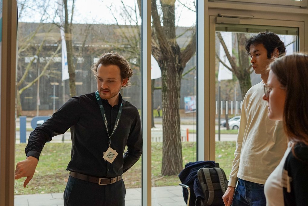 Matter to Life PhD candidate in dark shirt gesturing inside a glass-walled building, with backpacks nearby; trees visible outside through the windows.