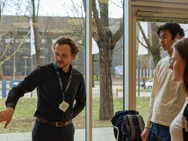 Matter to Life PhD candidate in dark shirt gesturing inside a glass-walled building, with backpacks nearby; trees visible outside through the windows.