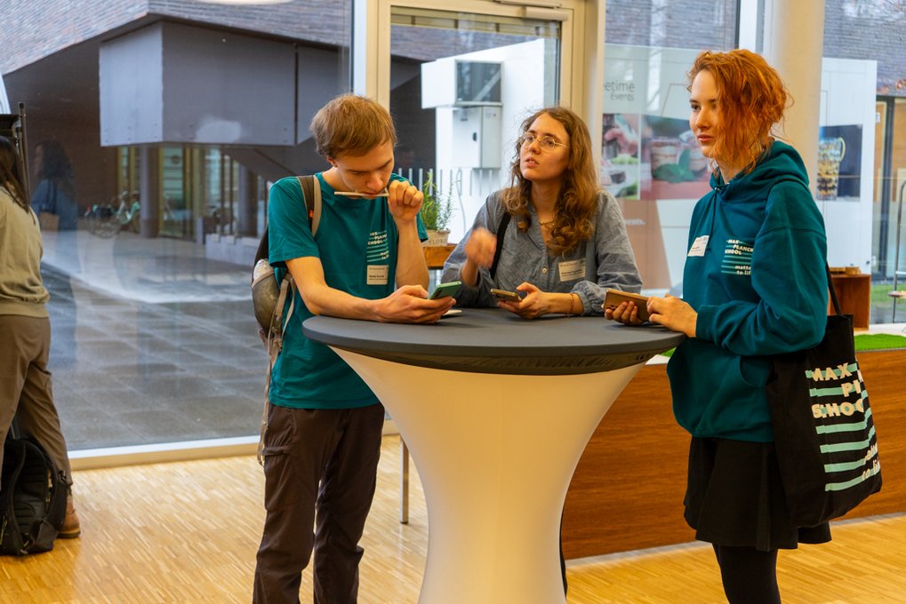 Three people are gathered around a high-top table in a contemporary indoor environment, holding smartphones and engaging in conversation. They are dressed casually, with one wearing a backpack and another carrying a tote bag.