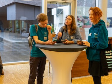 Three people are gathered around a high-top table in a contemporary indoor environment, holding smartphones and engaging in conversation. They are dressed casually, with one wearing a backpack and another carrying a tote bag.