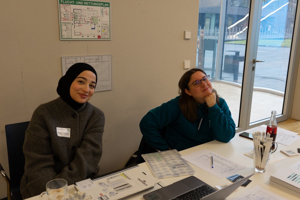 Two people sit at a table with various documents, a laptop, a bottle of soda, and writing utensils, positioned by a large window.