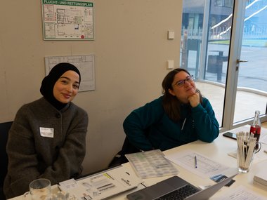 Two people sit at a table with various documents, a laptop, a bottle of soda, and writing utensils, positioned by a large window.