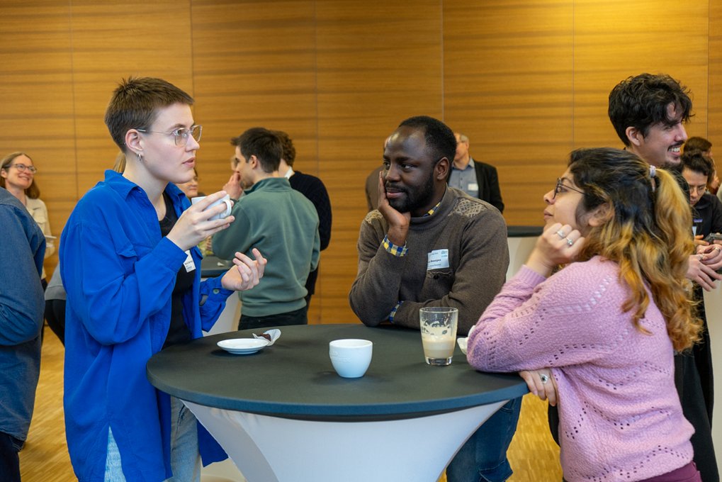 Individuals standing at a round table, having a conversation, holding cups, with a wooden wall in the background.