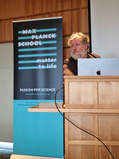 An indoor setting featuring a Max Planck School banner promoting science, with Philippe Bastiaens behind a wooden podium and an open laptop in the foreground.
