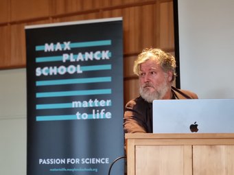 An indoor setting featuring a Max Planck School banner promoting science, with Philippe Bastiaens behind a wooden podium and an open laptop in the foreground.