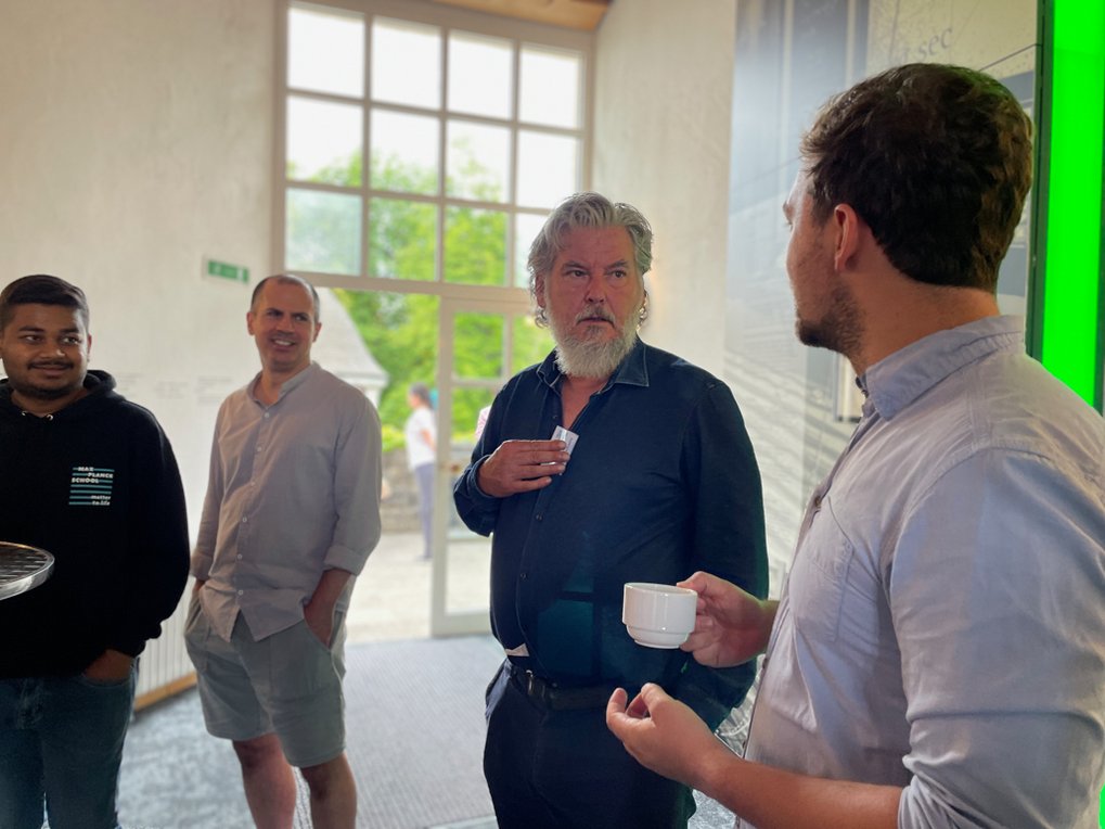 Four individuals stand in a well-lit room, engaging in conversation, with one person holding a white cup, near a bright green wall and large window.