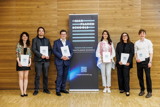 Seven graduates of the Max Planck Schools standing with their certificates in front of a Max Planck Schools banner – celebrating the successful completion of a scientific program