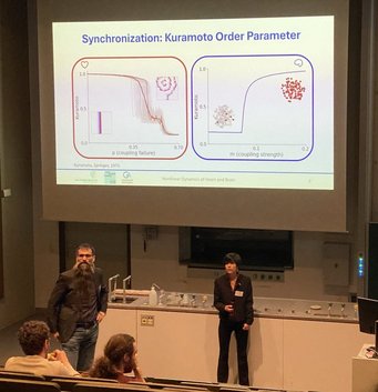 Irene presenting her research Academic presentation in a lecture hall focusing on the Kuramoto Order Parameter, featuring two graphs analyzing synchronization metrics related to coupling strength and failure, with attendees observing.