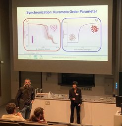 Academic presentation in a lecture hall focusing on the Kuramoto Order Parameter, featuring two graphs analyzing synchronization metrics related to coupling strength and failure, with attendees observing.