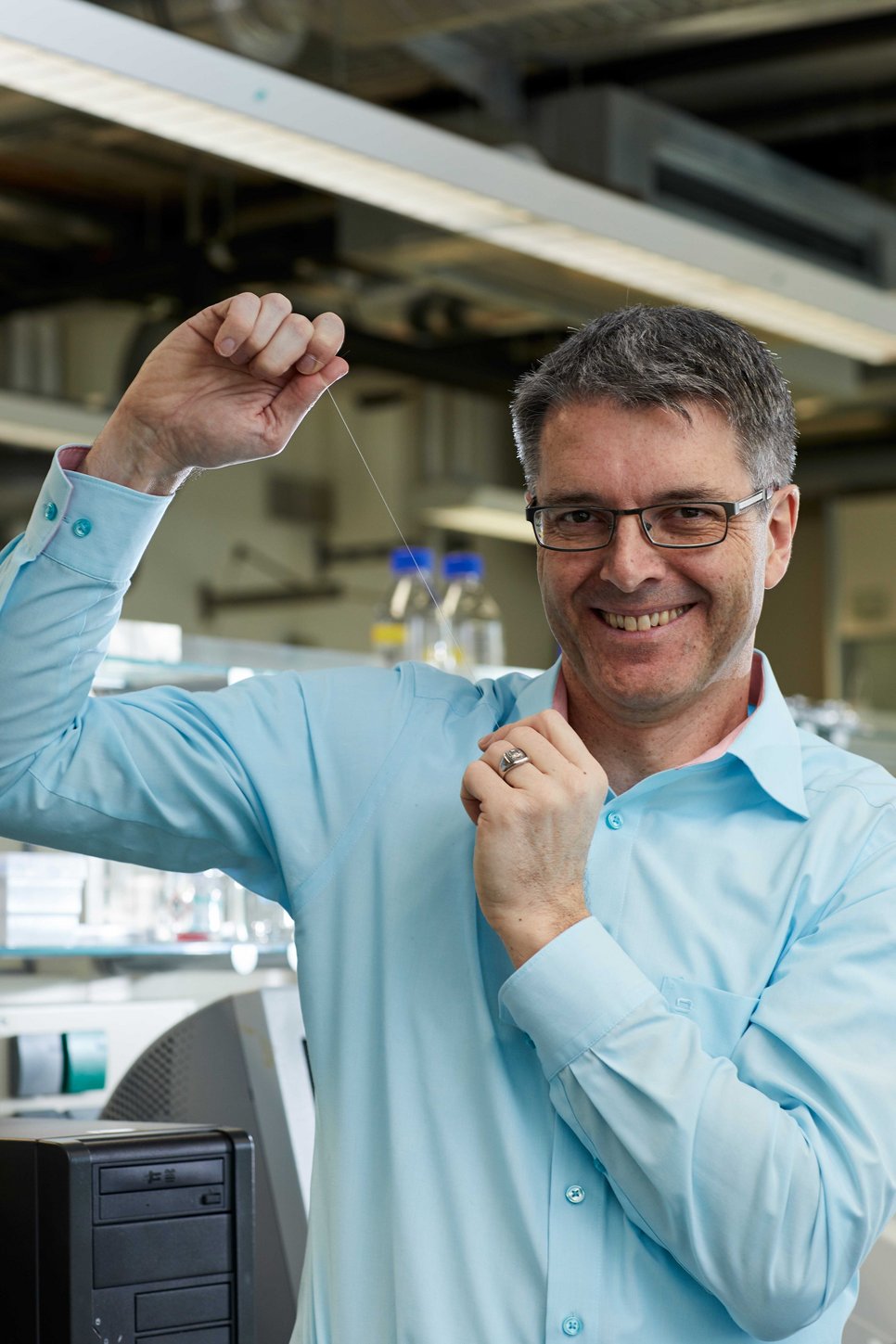 Matter to Life Fellow Thomas Scheibel, wearing a blue shirt, stands in a laboratory holding a thin, transparent thread between his fingers, surrounded by laboratory equipment.