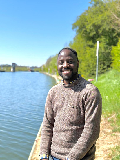 Deus standing by a riverbank on a sunny day with green trees and blue water in the background.