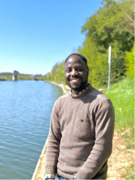 Deus standing by a riverbank on a sunny day with green trees and blue water in the background.