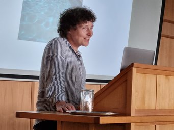 Petra Schwille connected past and present in a compelling keynote, setting an inspiring tone for the Symposium. Petra Schwille is standing at a wooden podium and speaking.