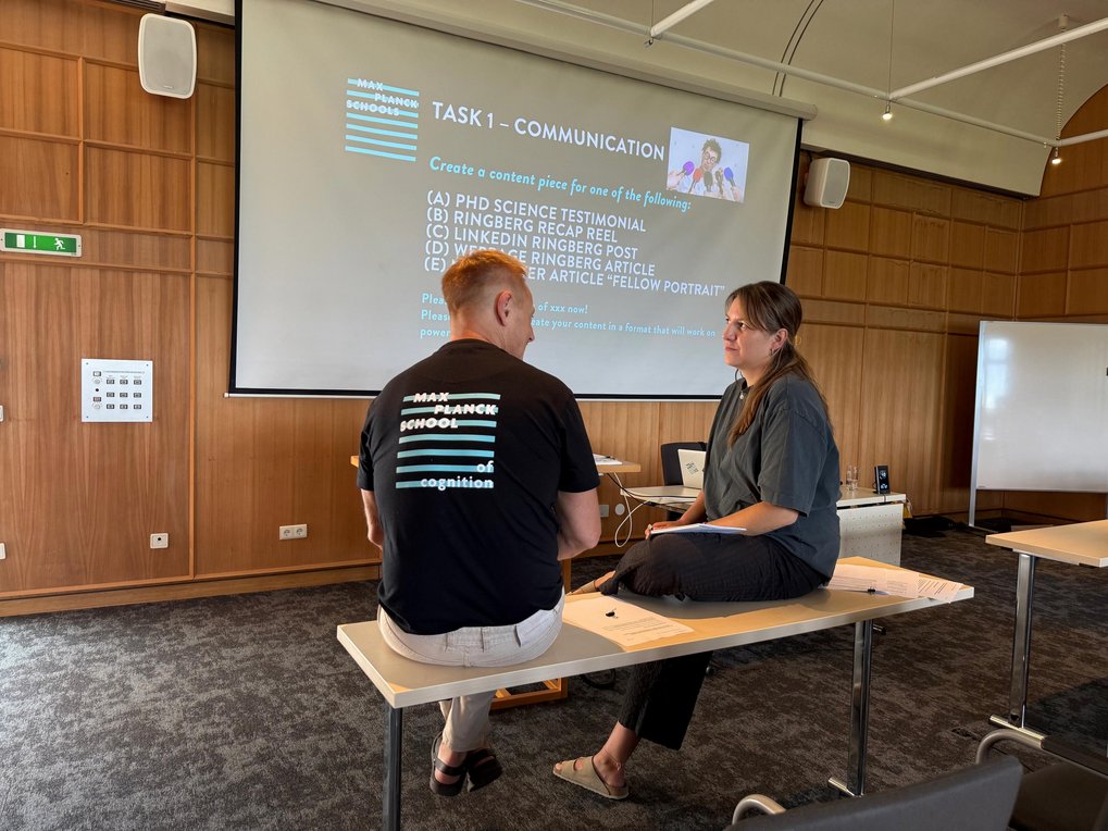 Discussing creative ways to communicate science—because research doesn’t make an impact if it stays in the lab. Two MPS coordinators sit on tables facing each other, discussing in front of a projector screen showing a slide titled "Task 1 – Communication." The slide lists options for creating science content.