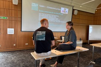 Discussing creative ways to communicate science—because research doesn’t make an impact if it stays in the lab. Two MPS coordinators sit on tables facing each other, discussing in front of a projector screen showing a slide titled "Task 1 – Communication." The slide lists options for creating science content.