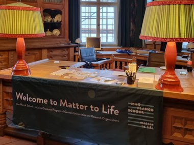 A wooden welcome desk in a historic room of the castle with large windows and ornate furniture. A banner on the desk reads “Welcome to Matter to Life” The desk holds name tags, pens, and informational materials.