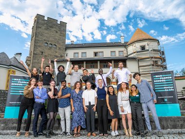 Group picture of the graduates, also using the castle as a backdrop. A few students in the front are hitting the thinking pose, a few in the back are raising their hands.