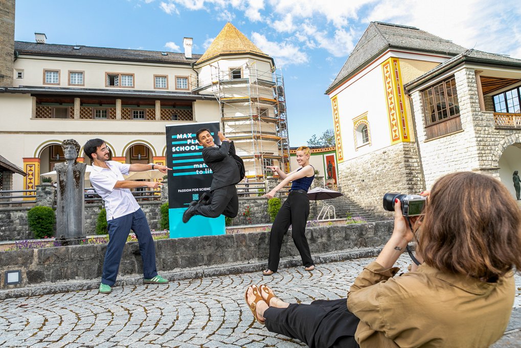 Three students pose playfully in front of a MPS MtL banner in front of the castle, while another student lies on the ground taking their photo with a camera. The student in the middle is mid-jump, while the two others stand on either side, pointing at them.