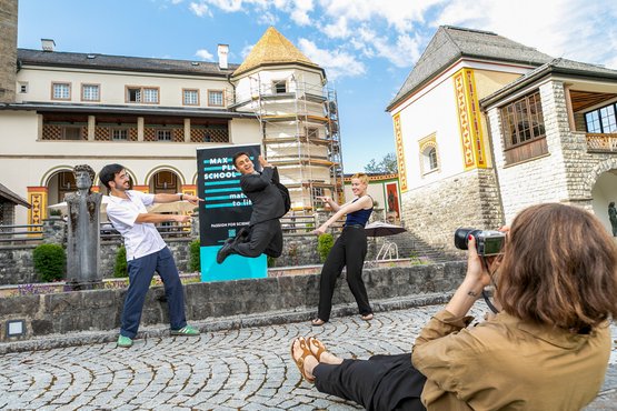Three students pose playfully in front of a MPS MtL banner in front of the castle, while another student lies on the ground taking their photo with a camera. The student in the middle is mid-jump, while the two others stand on either side, pointing at them.