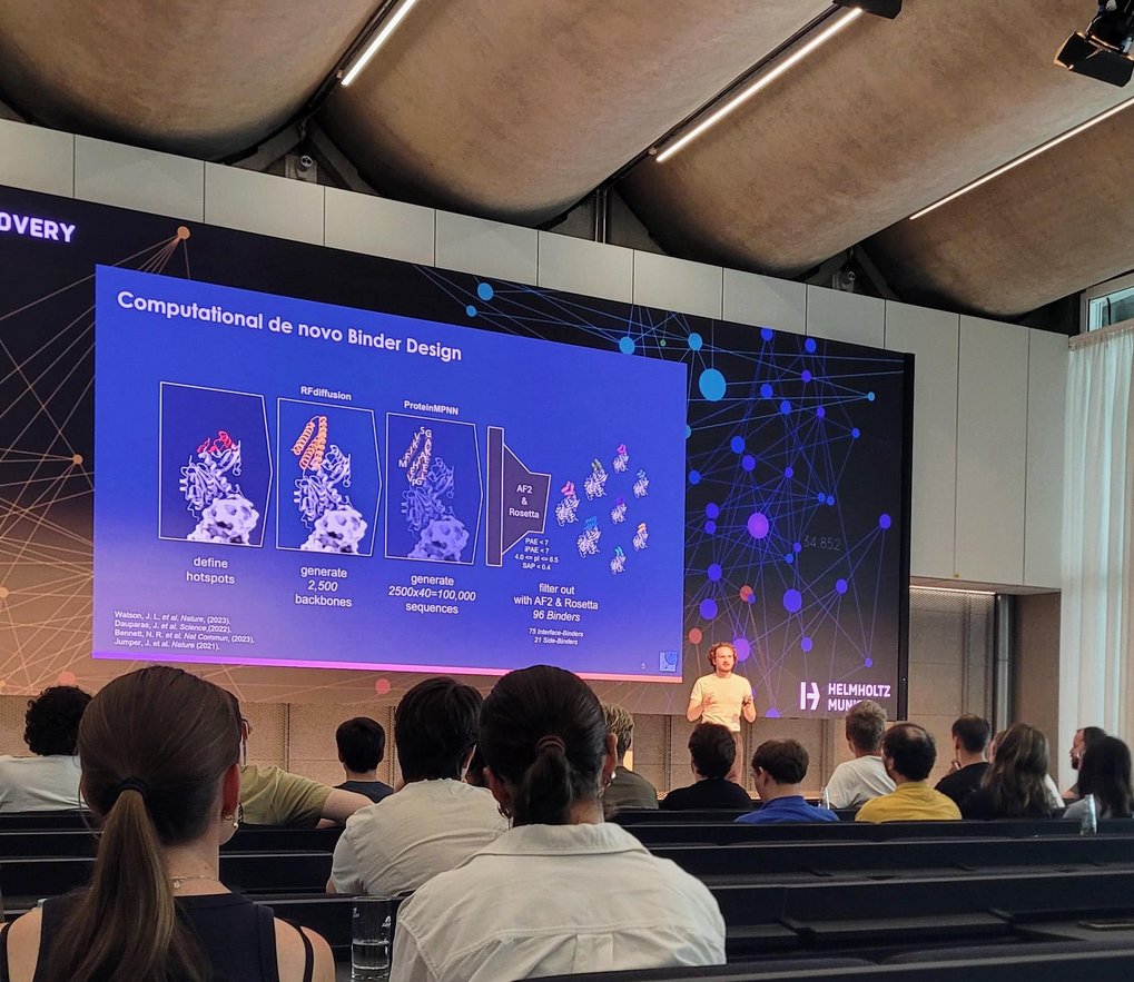 Andrey präsentiert vor einer großen Leinwand, auf der eine Folie mit dem Titel „Computational de novo Binder Design” mit Diagrammen von Proteinstrukturen und Text zu sehen ist, während das Publikum in einem Hörsaal zuhört.