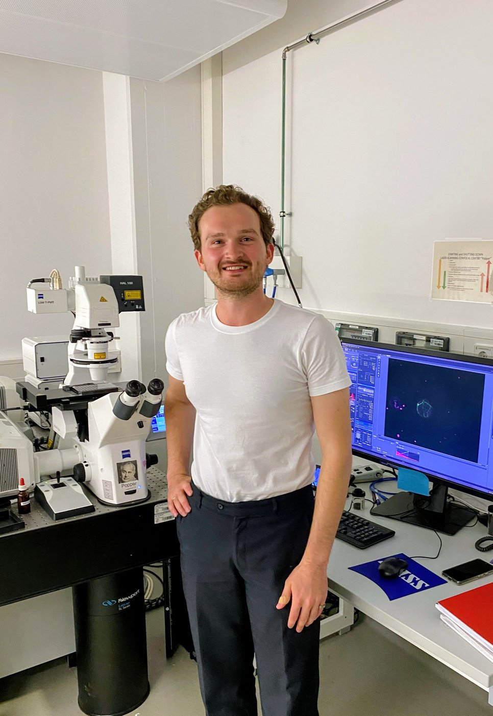 Andrey in einem Labor neben Hightech-Mikroskopiegeräten, Computermonitoren, auf denen wissenschaftliche Bilder und Datenanalysen zu sehen sind.