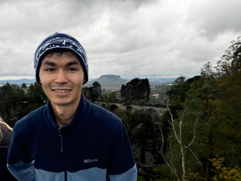 PhD candidate Mussa, with a stone bridge spans rocky cliffs with a cloudy sky in the background