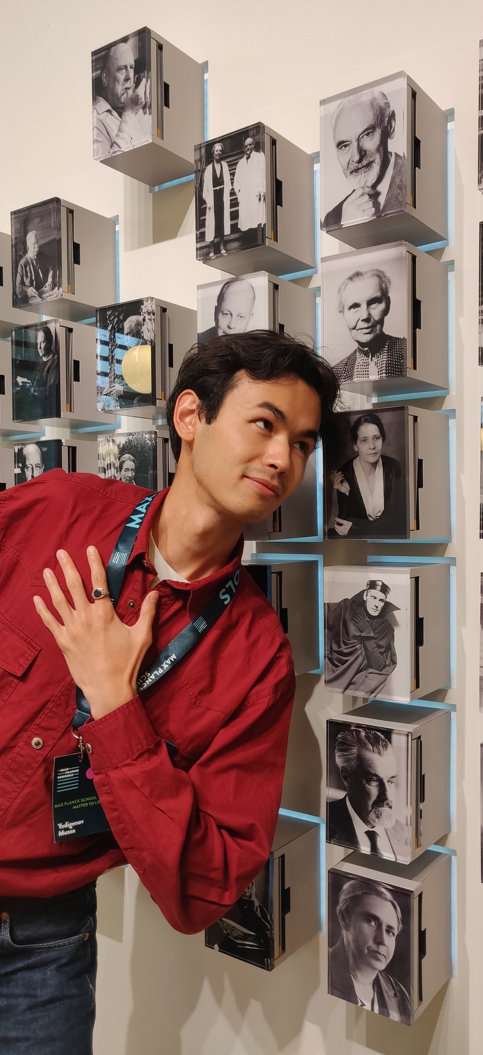 PhD candidate Mussa in a red shirt with a lanyard stands beside a wall featuring black-and-white photographs in box frames with soft lighting.