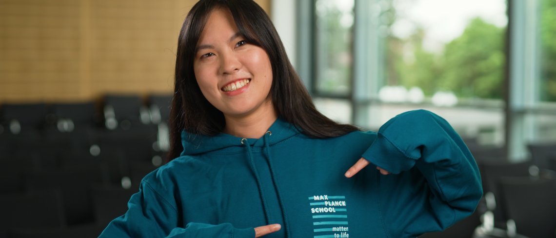 Lächelnde junge Frau in einem türkisfarbenen Kapuzenpullover, die auf das Logo auf ihrem Sweatshirt zeigt, stehend in einem modernen, gut beleuchteten Auditorium mit leeren Stühlen im Hintergrund.