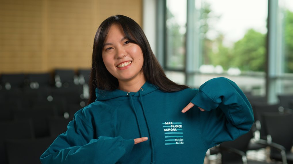 Lächelnde junge Frau in einem türkisfarbenen Kapuzenpullover, die auf das Logo auf ihrem Sweatshirt zeigt, stehend in einem modernen, gut beleuchteten Auditorium mit leeren Stühlen im Hintergrund.