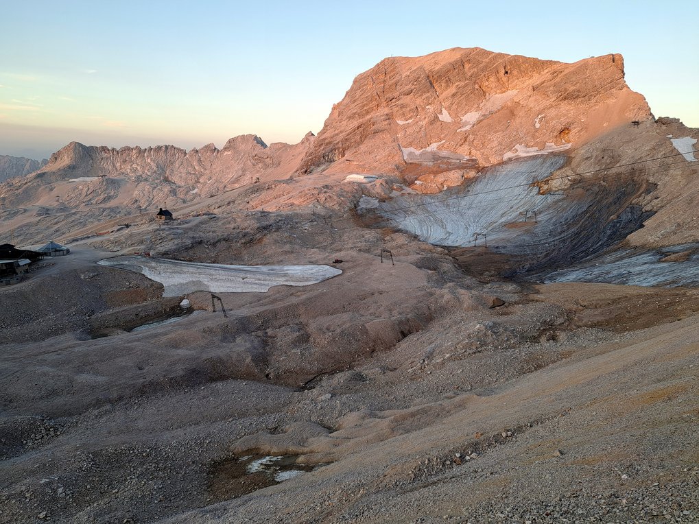 Mountain peak illuminated by sunlight, surrounded by rocky terrain and isolated snow remnants.