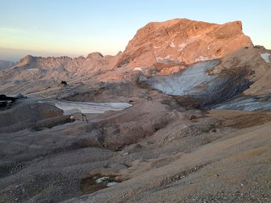 Mountain peak illuminated by sunlight, surrounded by rocky terrain and isolated snow remnants.