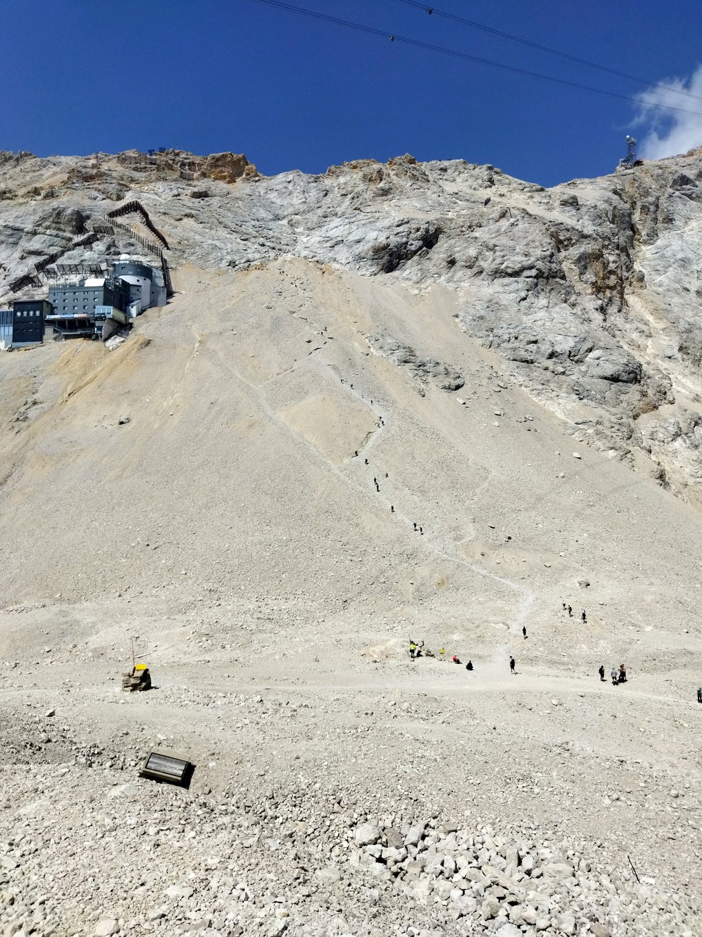 Steep rocky mountain slope with hikers ascending a narrow path, building visible on the left.
