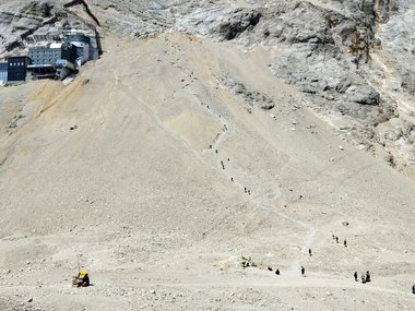 Steep rocky mountain slope with hikers ascending a narrow path, building visible on the left.