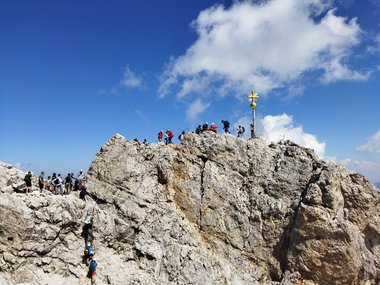 Groups of people standing on a jagged mountain peak featuring a prominent gold cross against a bright sky.