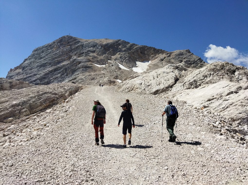 Four Symposium attendees climb a steep, rocky path toward a mountain summit, surrounded by rugged terrain and patches of snow.