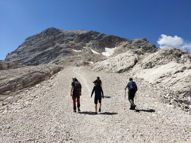 Four Symposium attendees climb a steep, rocky path toward a mountain summit, surrounded by rugged terrain and patches of snow.