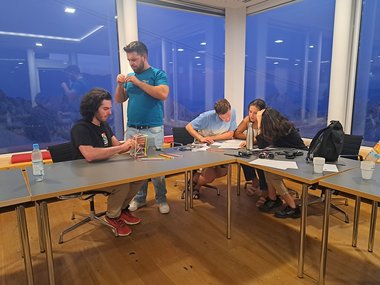 Five PhD candidates engaged in an activity, surrounded by papers, pencils, and a bottle on tables.