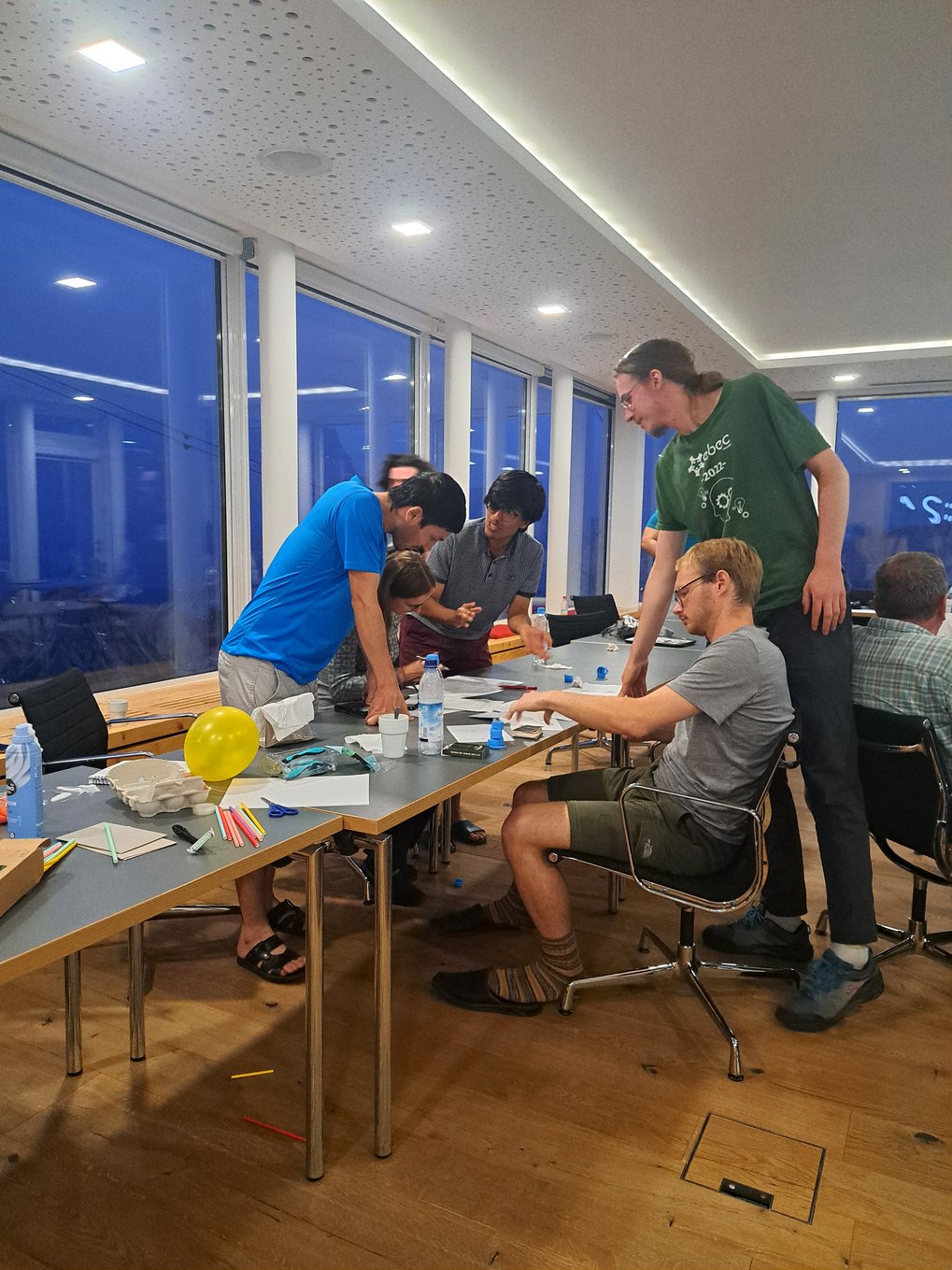 Several PhD candidates are engaged in a collaborative activity at a table filled with various craft supplies, including balloons, straws, and paper, in a spacious and modern room with large windows.