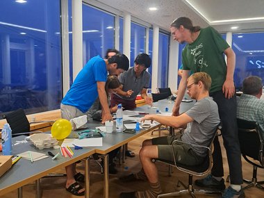 Several PhD candidates are engaged in a collaborative activity at a table filled with various craft supplies, including balloons, straws, and paper, in a spacious and modern room with large windows.