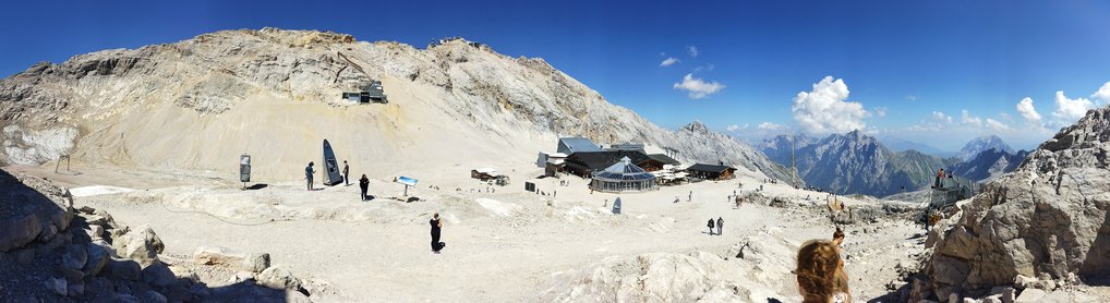 Panorama of a rocky alpine area with visitors walking near a mountain hut and distant peaks in view.