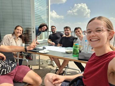 A group of Matter to Life PhD candidates are sitting at a table on a terrace with drinks and papers. In the background, a few clouds can be seen in the sky.