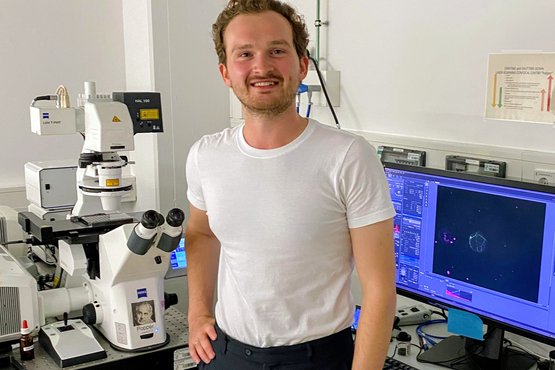 Andrey in einem Labor neben Hightech-Mikroskopiegeräten, Computermonitoren, auf denen wissenschaftliche Bilder und Datenanalysen zu sehen sind.