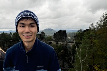 PhD candidate Mussa, with a stone bridge spans rocky cliffs with a cloudy sky in the background