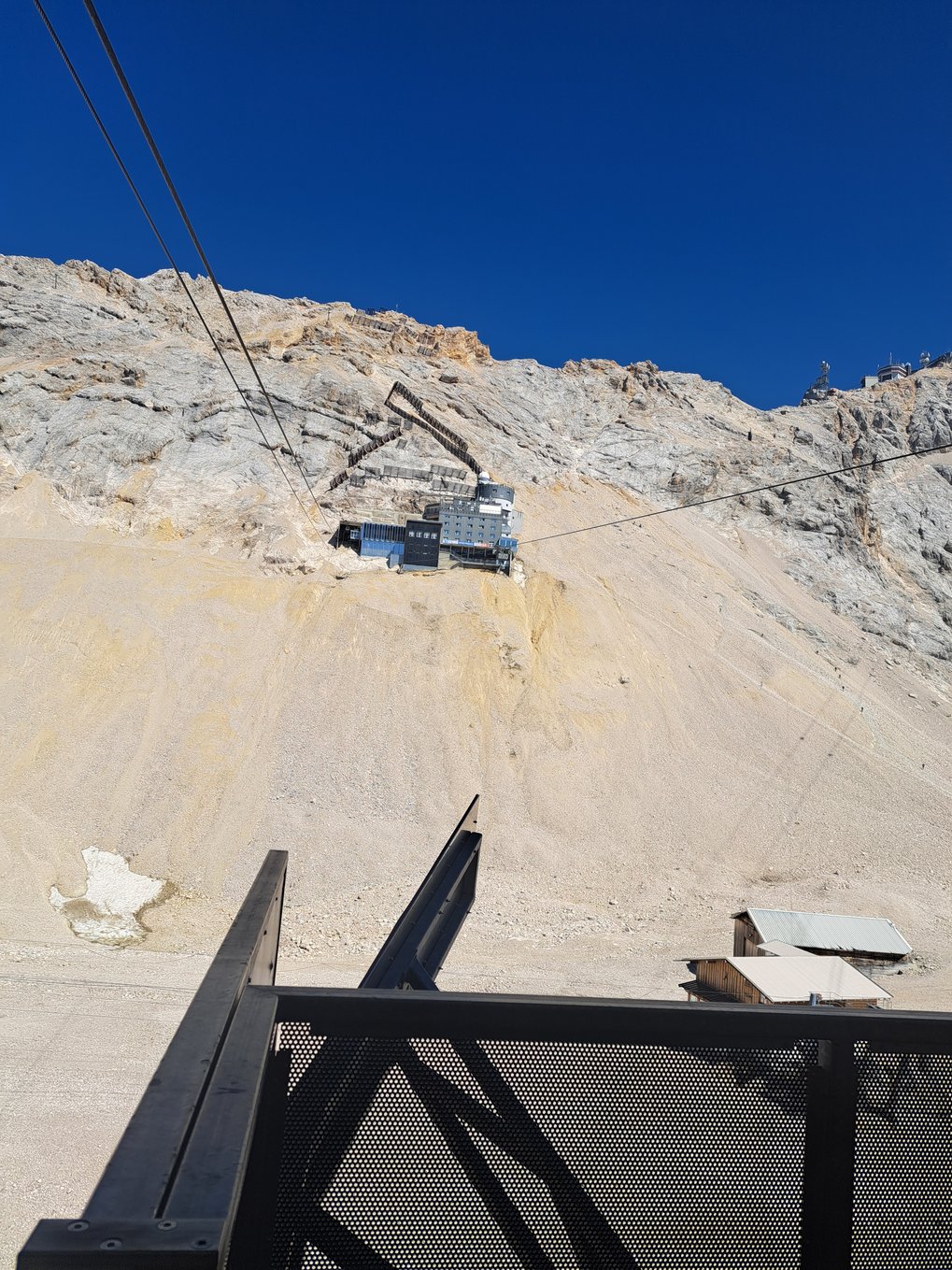 View of a cable car ascending a rocky mountain slope toward the Schneefernerhaus research station on the Zugspitze under a clear blue sky.