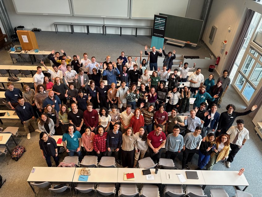 The whole Matter to Life Fall Days crew—physicists, chemists, biologists, and everything in between. “Say Science”! A diverse group of individuals is gathered in a lecture hall. They are smiling and waving at the camera. Desks are arranged in rows, and a presentation area with a blackboard and a banner is visible in the background.