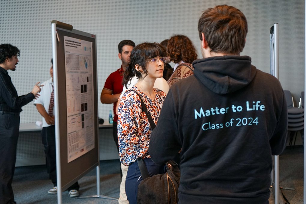 Attendees engage in conversation near a scientific poster; one wears a hoodie labeled "Matter to Life, Class of 2024."