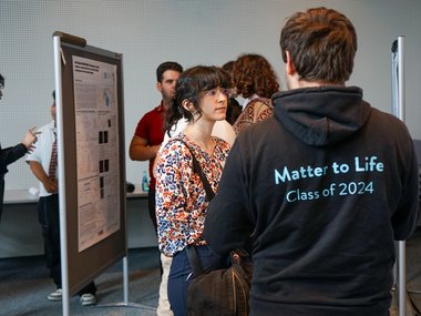 Attendees engage in conversation near a scientific poster; one wears a hoodie labeled "Matter to Life, Class of 2024."
