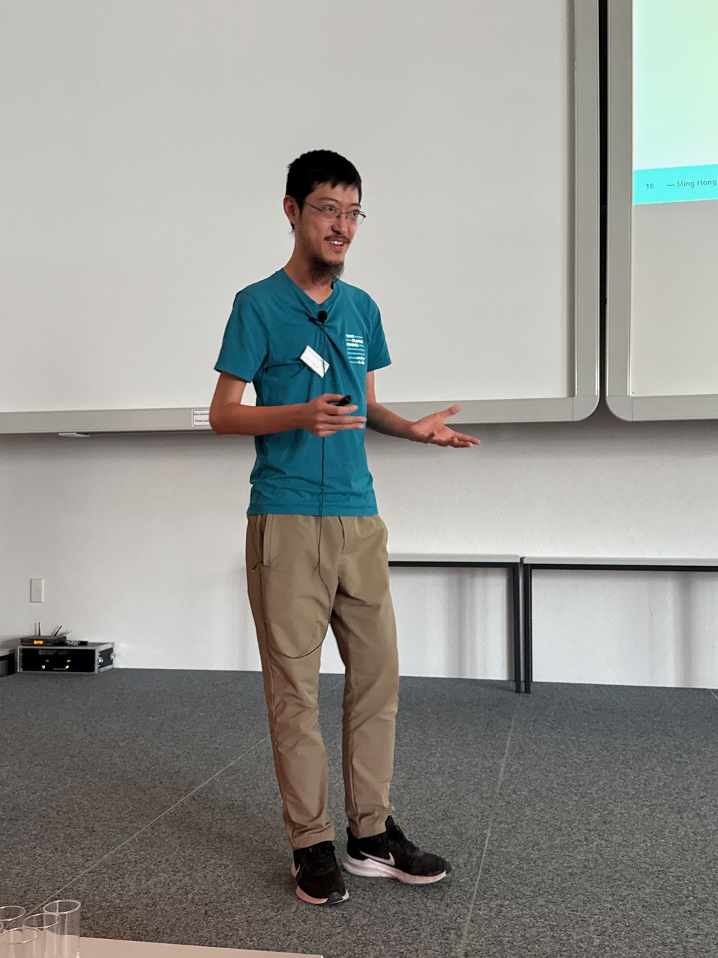 PhD candidate James in casual attire presenting a topic on stage, gesturing with hands.