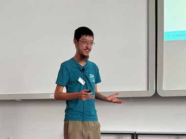 PhD candidate James in casual attire presenting a topic on stage, gesturing with hands.