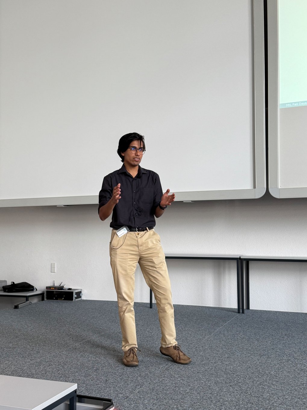 Matter to Life PhD candidate Pratulya in a dark shirt and khaki pants gestures on a stage, with a large projection screen behind them.